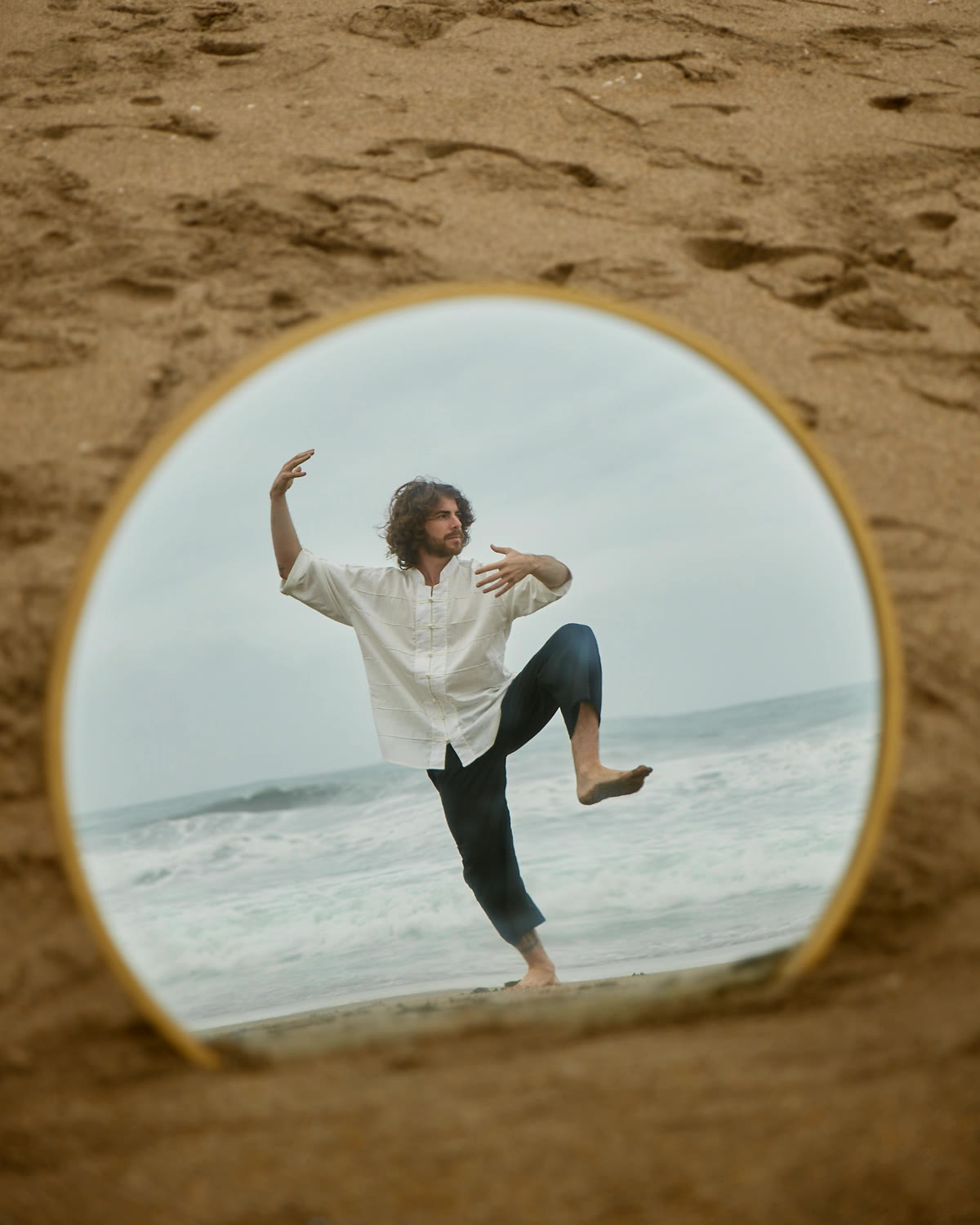 Rodri en una postura de Taichi frente al mar, reflejado en un espejo circular sobre la arena
